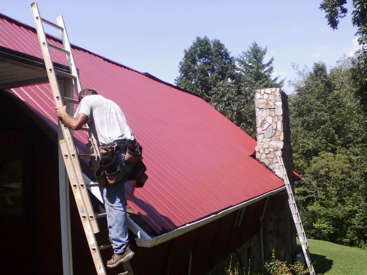 Roofer on a ladder beside a red metal roof and stone chimney — Soffit & Fascia Repair by Roofing Brothers in Turbeville